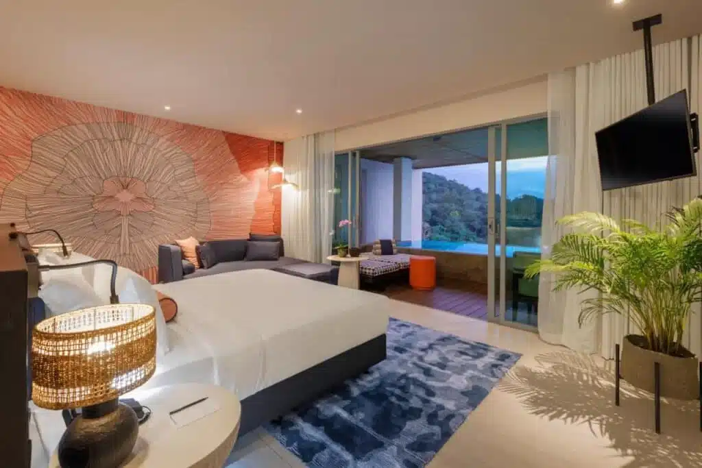 A hotel room with a modern design features a large bed, a cozy lounge area, and a wall-mounted TV. The room has a vibrant floral accent wall, contemporary furniture, and sliding glass doors leading to a balcony with outdoor seating and scenic views of the mountains and blue sky—perfect for those seeking hotels in Phuket.