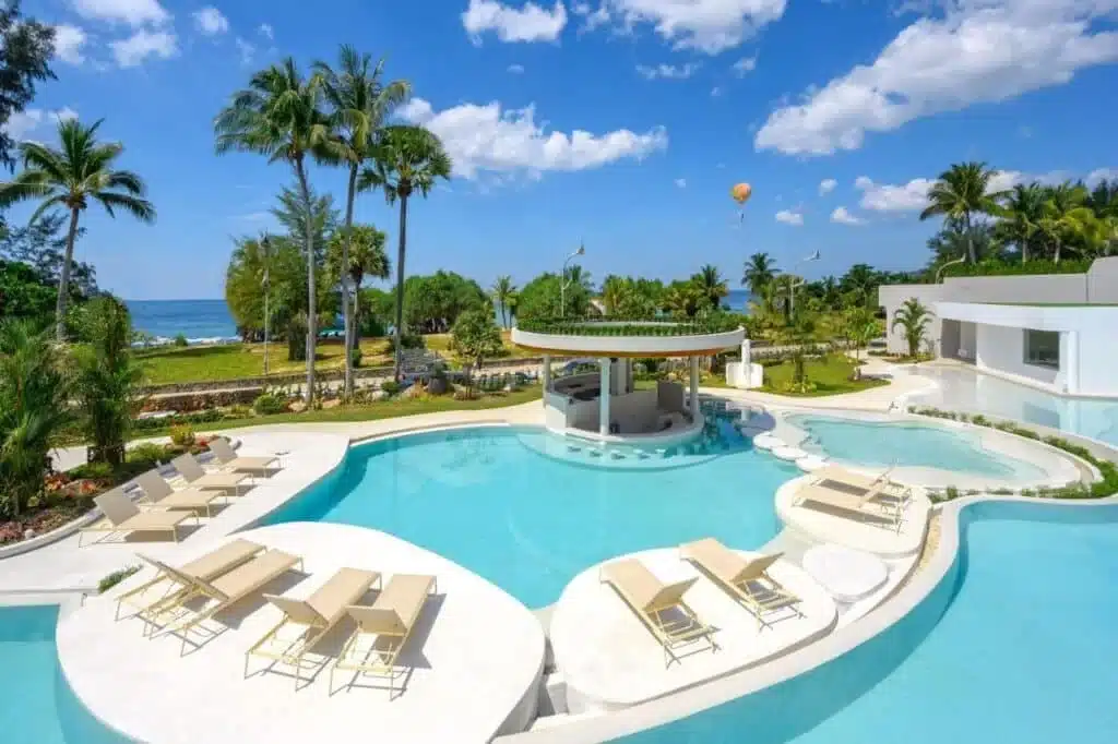 A luxurious outdoor pool area with a swim-up bar, surrounded by lounge chairs and palm trees, reminiscent of the finest hotels in Phuket. The pool has multiple levels and curved edges. In the background, there's a view of the ocean, a hot air balloon in the sky, and lush greenery under a partly cloudy blue sky.