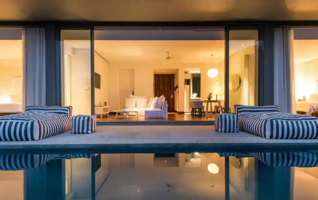 A modern, luxurious poolside living area is seen at night. Two striped loungers flank an inviting pool, while warm, yellow lighting fills the interior of the adjacent house. Large sliding glass doors open to reveal a stylish, well-lit living room with contemporary furnishings and decor, reminiscent of top hotels in Phuket.