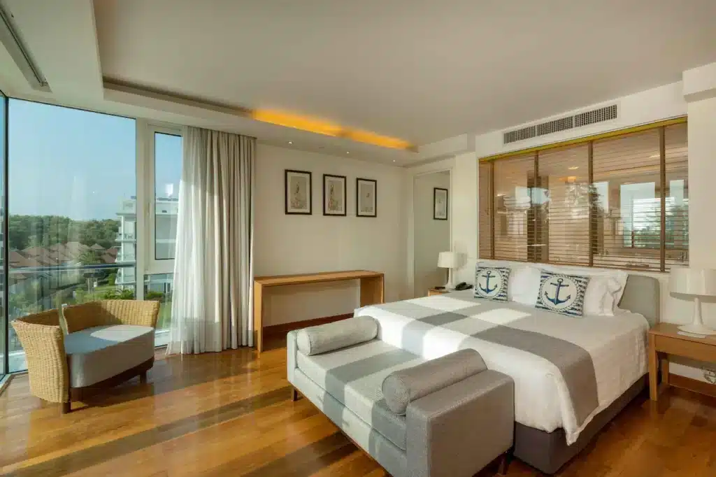 A spacious bedroom with a large window letting in natural light. It features a king-sized bed with white and gray striped bedding and anchor-themed pillows, a wicker chair and small table by the window, a large desk, and framed nautical artwork above the bed. The room has wooden flooring, reminiscent of top hotels in Phuket.