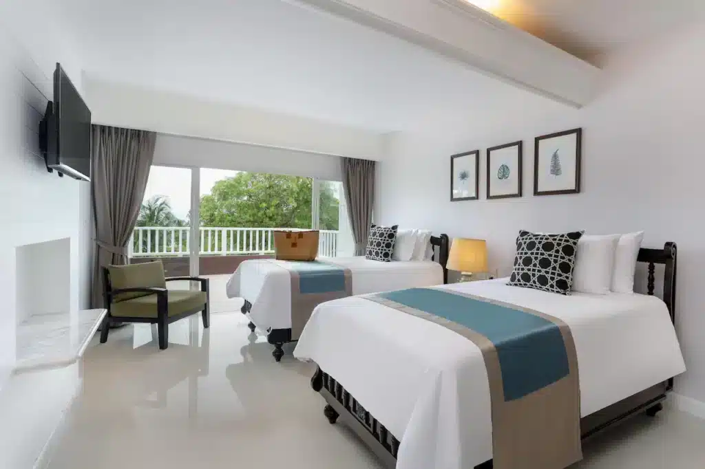 A modern hotel room, typical of hotels in Phuket, features two neatly made beds with white linens and teal runners. Above the beds are three framed artworks. A wall-mounted TV faces the beds, and a comfortable armchair sits next to a large window with a view of trees. A bedside lamp emits a warm glow.