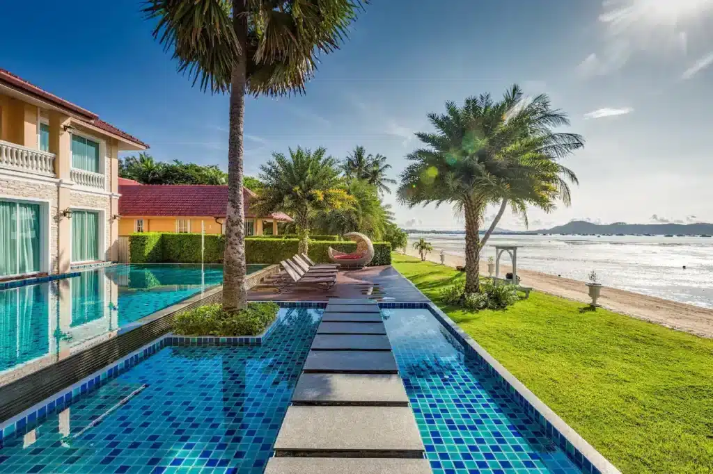 A serene beachfront resort in Phuket features a pool with a stepping stone path leading to a row of sun loungers under a palm tree. A nearby large round daybed offers a cozy seating spot. The resort building has red roofs and colorful exteriors, and the calm ocean is visible under a clear sky.