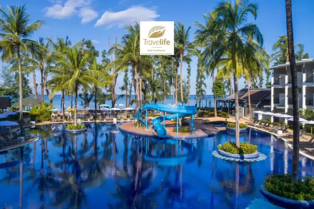 A luxurious resort pool with a central blue water slide encircled by lush tropical trees and palm trees. Lounge chairs and umbrellas line the poolside. The sea is visible in the background. The "Travelife Gold Certified" logo is prominently displayed, showcasing one of the finest hotels in Phuket.