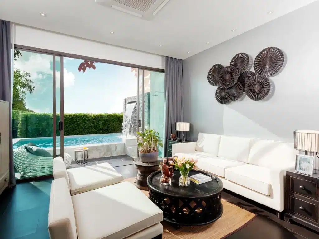 A modern living room featuring white sofas, a round black coffee table, and wall art with circular motifs. A floor-to-ceiling glass door opens to the pool and garden area with a lush hedge. Natural light illuminates the space, and various plants add greenery—perfect for those seeking Private Pool Villas in Phuket.