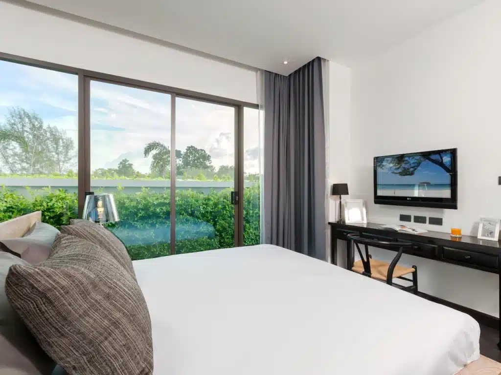 A modern bedroom in a Phuket pool villa with a large window on the left featuring sliding glass doors that open to a garden view. The room has a neatly made bed with gray and white bedding, a dark wooden desk with a lamp, framed pictures, and a wall-mounted flat-screen TV adjacent to light gray curtains.