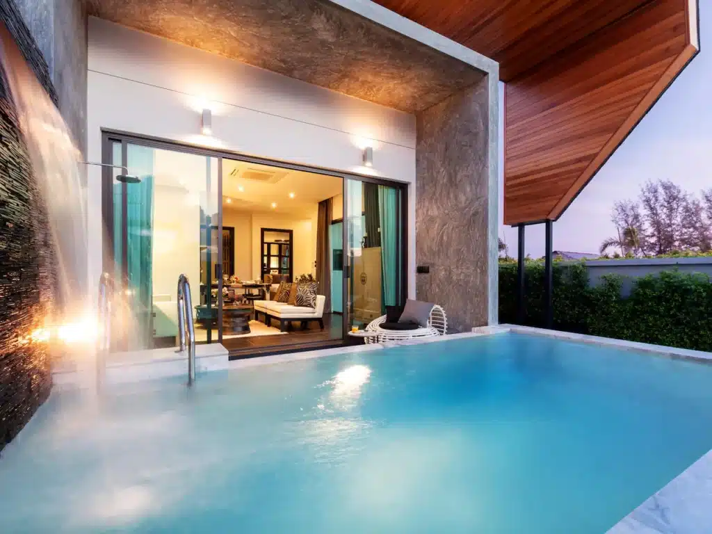 A luxurious poolside scene featuring a modern home with large sliding glass doors opening to an elegantly furnished interior. The Phuket pool villa, accentuated by a cascading waterfall on the left, has a nearby patio area with outdoor seating. The house exterior has wood-panel accents and lush greenery surrounds the area.