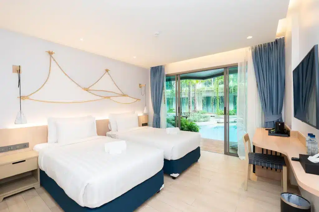 A brightly lit hotel room at one of the charming hotels in Phuket features two neatly made twin beds with white linens and blue skirts. The room has a large glass sliding door leading to a garden and pool area outside. A wooden desk with a chair is placed against the wall, next to a flat-screen TV mounted above.