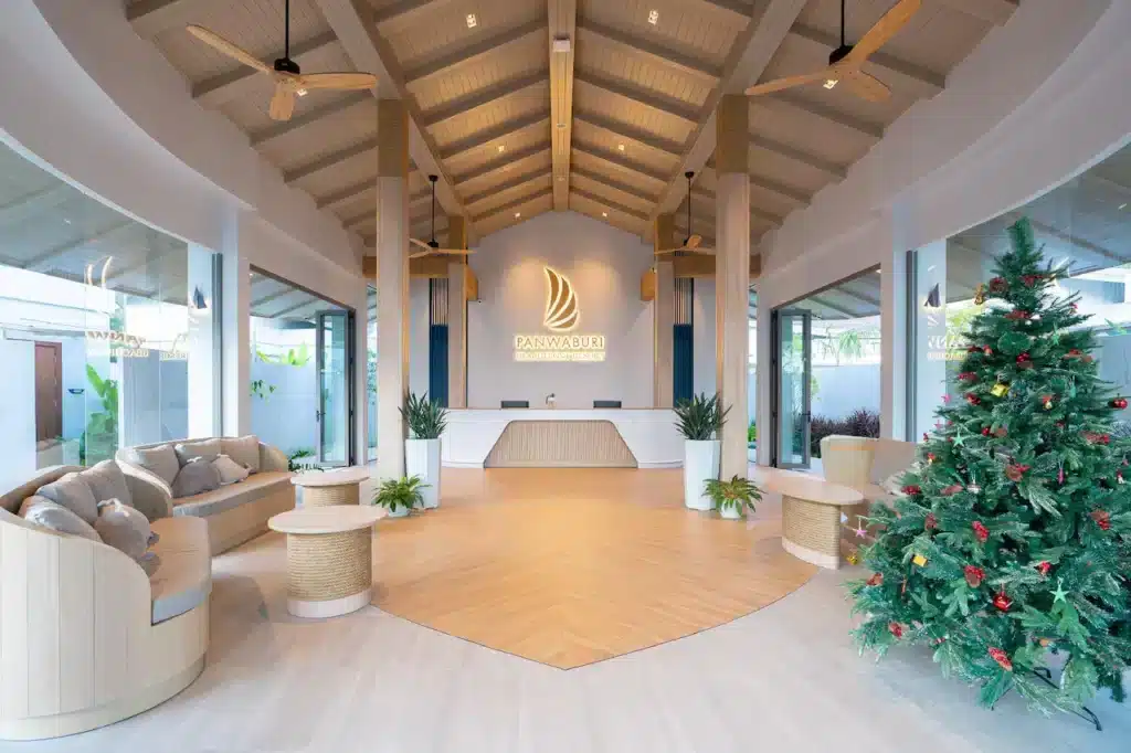 A spacious, modern hotel lobby with high, wooden ceilings and ceiling fans. The room features a minimalist design with neutral colors. There is a large reception desk in the center, flanked by sofas and potted plants. A decorated Christmas tree stands on the right side of the image, embodying the elegance found in hotels in Phuket.