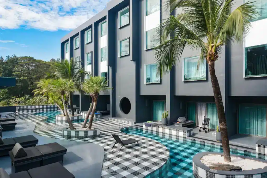 A modern hotel courtyard reminiscent of the best hotels in Phuket, featuring a curvy, checkered black-and-white tiled pool surrounded by palm trees and cushioned lounge chairs. The three-story building has large windows and a sleek, dark gray exterior, all under a partly cloudy sky creating a tropical, relaxing atmosphere.