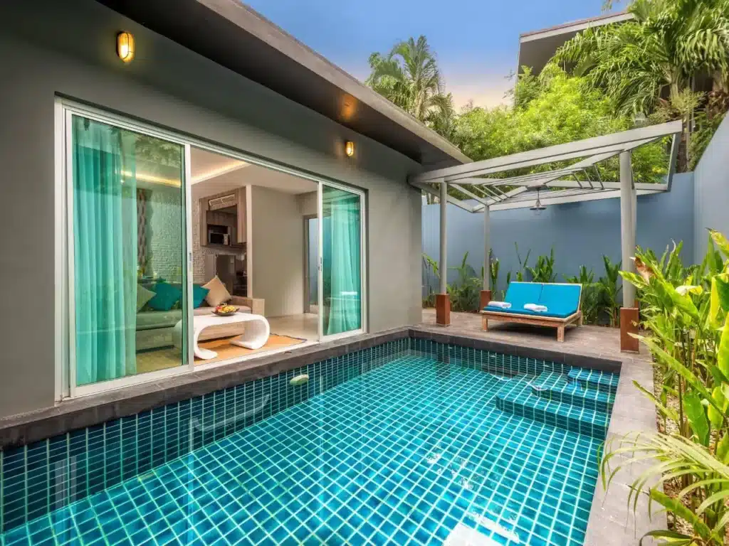 A modern Phuket pool villa features a small, rectangular swimming pool with a blue tile design. Adjacent to the pool is a glass sliding door leading into a living area with teal curtains. A shaded patio area with two blue cushioned lounge chairs and green plants surround the space.