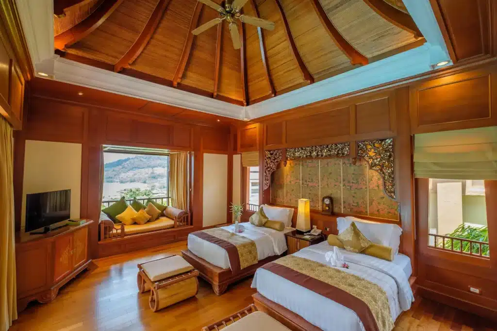 A tropical-themed room in our Phuket pool villa features two single beds with white linens and golden accents. The wooden ceiling and walls add warmth, and a large window opens to a scenic view. It has a ceiling fan, TV, seating area with cushions, and a detailed headboard. Decor is elegant and inviting.