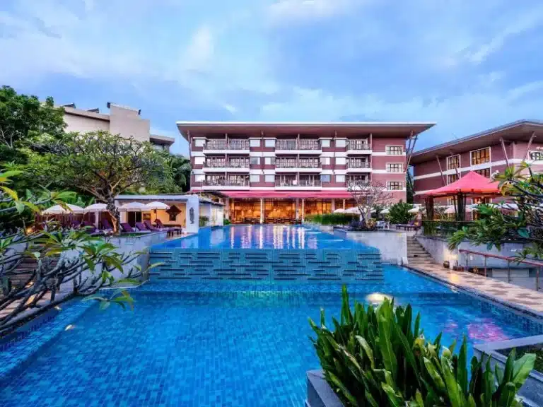 A luxurious swimming pool lined with greenery leads to a modern, multi-story hotel building. Lounge chairs and shaded canopies are arranged around the pool area, with trees and plants adding a tropical touch. Private Pool Villas in Phuket enhance the ambiance as balcony lights glow warmly under a partly cloudy sky.