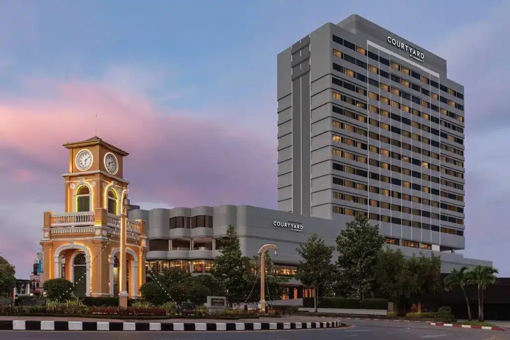 A modern high-rise hotel building labeled "Courtyard" stands tall against a pink and blue sky. To its left stands a yellow, classical clock tower with arched windows and a clock face. Both structures are surrounded by landscaped greenery and a road with striped pedestrian crossings in the foreground, reminiscent of hotels in Phuket.