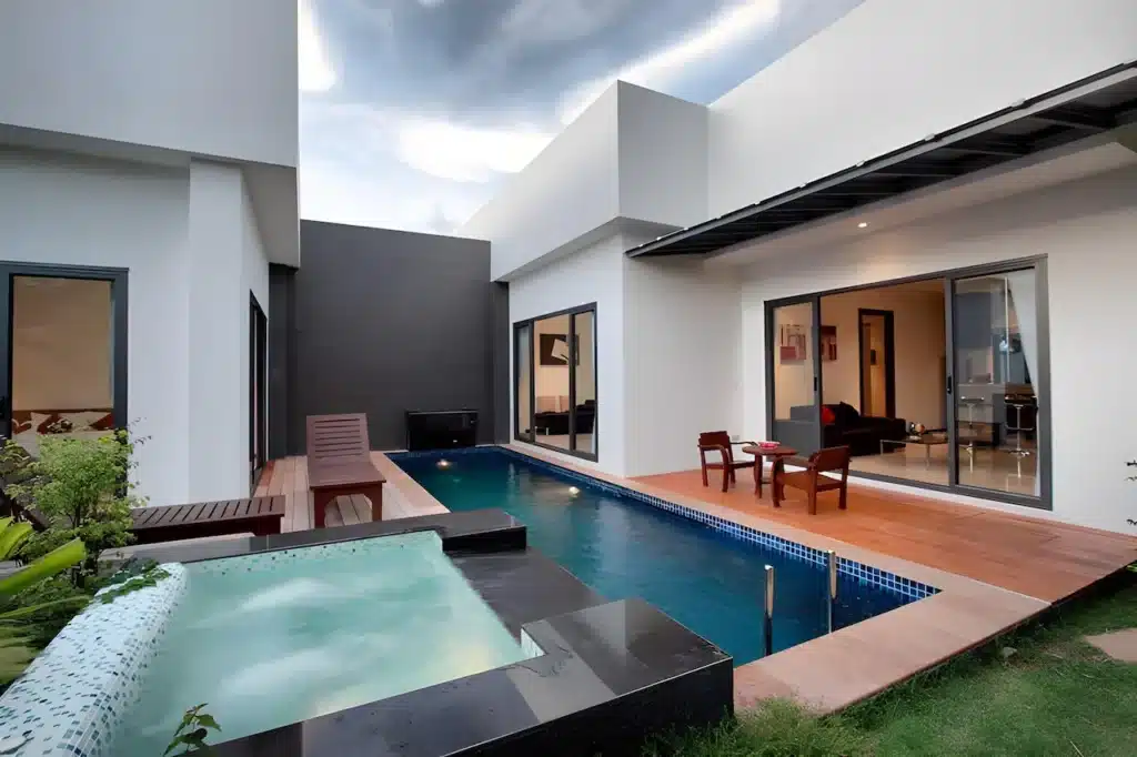 A modern residential outdoor space in a Phuket pool villa features a rectangular pool with a wooden deck and adjacent hot tub. The area is surrounded by clean, geometric white walls with large glass sliding doors leading to the interior. Minimalistic wooden chairs and a table are set on the deck next to the pool.