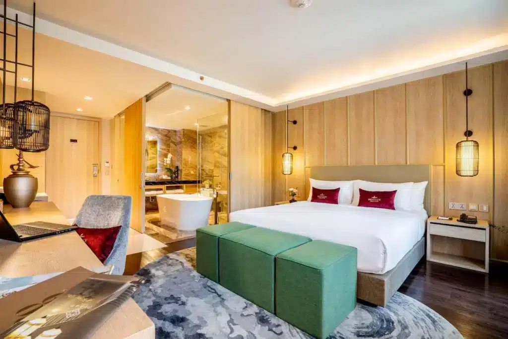 A modern hotel room featuring a spacious bed with white linens and red accent pillows. There are two green ottomans at the foot of the bed and hanging pendant lights on either side. The room opens up to a luxurious bathroom with a freestanding bathtub, glass walls, and elegant marble finishes, perfect for unwinding after exploring Natural Attractions in Bangkok.