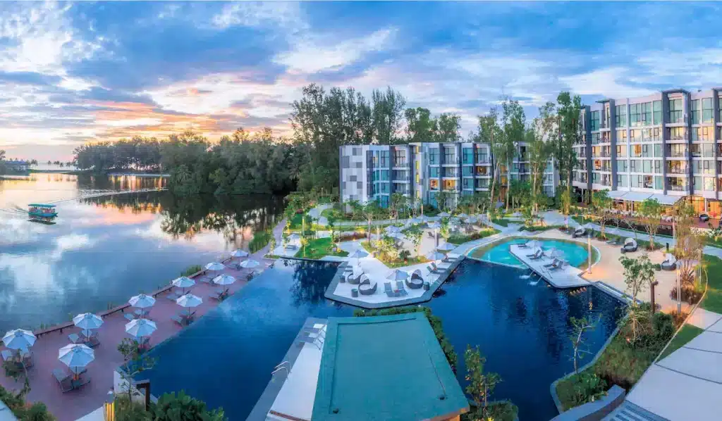 A scenic view of a luxurious waterfront resort features modern buildings with large windows, a spacious outdoor pool area with sun loungers, umbrellas, and a circular pool. Lush greenery and trees surround the area. A serene lake mirrors the vibrant sky during sunset—a gem among hotels in Phuket.