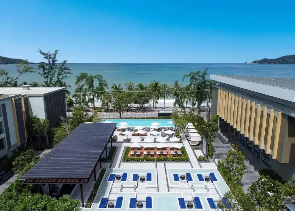 Aerial view of a tropical beach resort with a large swimming pool surrounded by lounge chairs and umbrellas. Modern buildings border the pool area. Beyond the resort, lush palm trees line the beach with calm, clear blue ocean water stretching out to the horizon under a bright blue sky—one of many stunning hotels in Phuket.