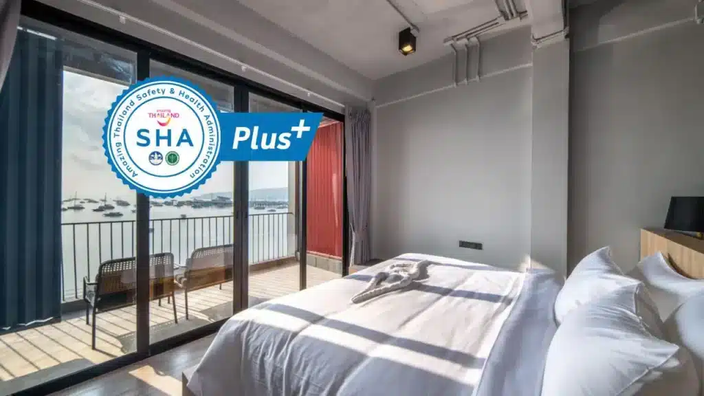 A modern hotel room with a large bed adorned with white linens. Sunshine filters through large glass doors, leading to a balcony with chairs overlooking a serene seascape with boats. Among the finest hotels in Phuket, it boasts a "SHA Plus+" certification badge on the left, indicating health and safety compliance.