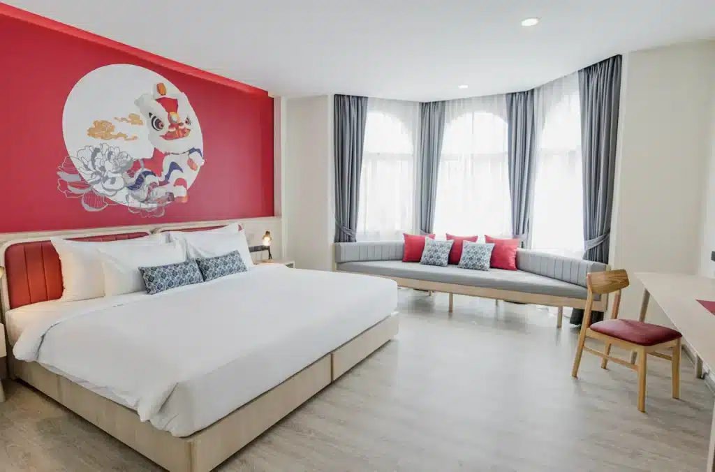 A brightly lit modern bedroom featuring a large bed with white linens and red accents, set against a striking red wall adorned with a decorative graphic of a koi fish. With references from Phuket travel guides, the seating area in front of two tall windows boasts cushions in matching red and white tones.