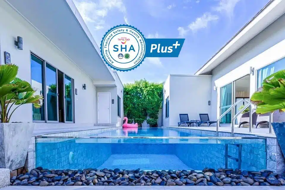 A serene outdoor pool area at a Phuket pool villa featuring clear blue water surrounded by white buildings. There is a pink flamingo pool float in the water. Wooden deck chairs are placed next to the pool, and plants in planters provide greenery. The SHA Plus+ certification emblem is displayed in the upper portion of the image.