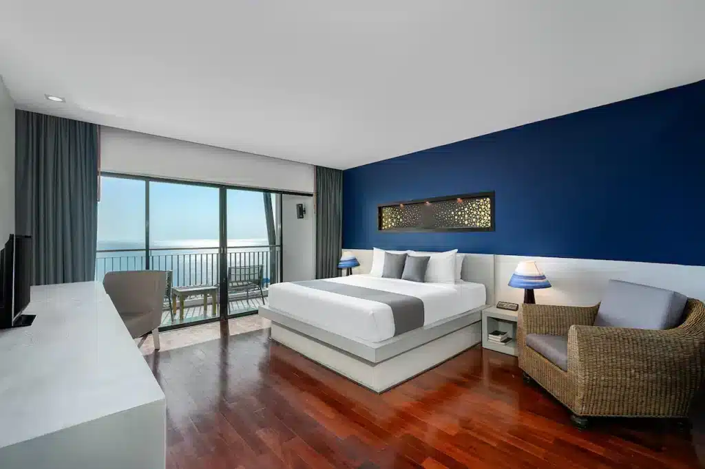 A modern hotel room featuring a large bed with a blue accent wall behind it. The room includes two bedside tables with lamps, a wicker armchair with a cushion, and a flat-screen TV on a white desk. Floor-to-ceiling windows offer an ocean view and balcony with table and chairs, perfect for those seeking hotels in Phuket.