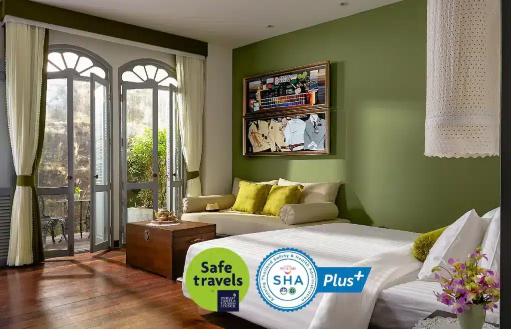 A bright, cozy room features a large bed with green and white pillows, and a green accent wall adorned with a framed picture. Sunlight streams through arched double doors leading to a balcony. A wooden trunk sits at the foot of the bed, alongside certification logos for "Safe travels" and "SHA Plus+," recommended in every Phuket travel guide.