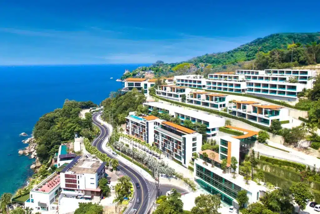 Aerial view of a luxurious coastal resort with multiple modern white and glass buildings on a hillside. The resort overlooks a scenic ocean, with a winding coastal road and lush green forest. Among the finest hotels in Phuket, the clear blue sky enhances the overall tranquility and beauty of the landscape.