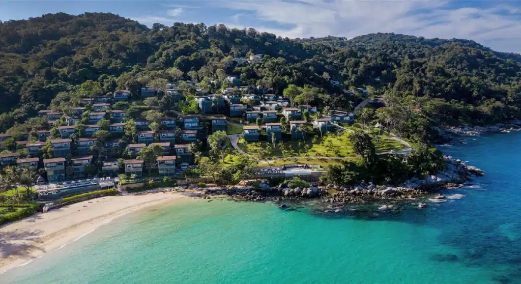 Aerial view of a coastal resort area with numerous buildings nestled among lush greenery on a hillside. The buildings, including luxurious Phuket pool villas, overlook a serene, sandy beach with turquoise waters. The surrounding landscape features dense forested areas and rocky shoreline, with clear skies above.