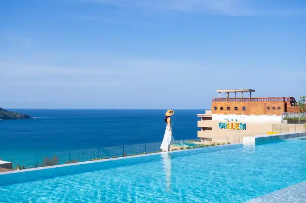 A woman in a white dress and sunhat stands at the edge of an infinity pool, overlooking the deep blue sea and vibrant blue sky. In the background, a building with a brown deck and colorful signage sits against a distant green hillside—an ideal scene from one of the finest hotels in Phuket.