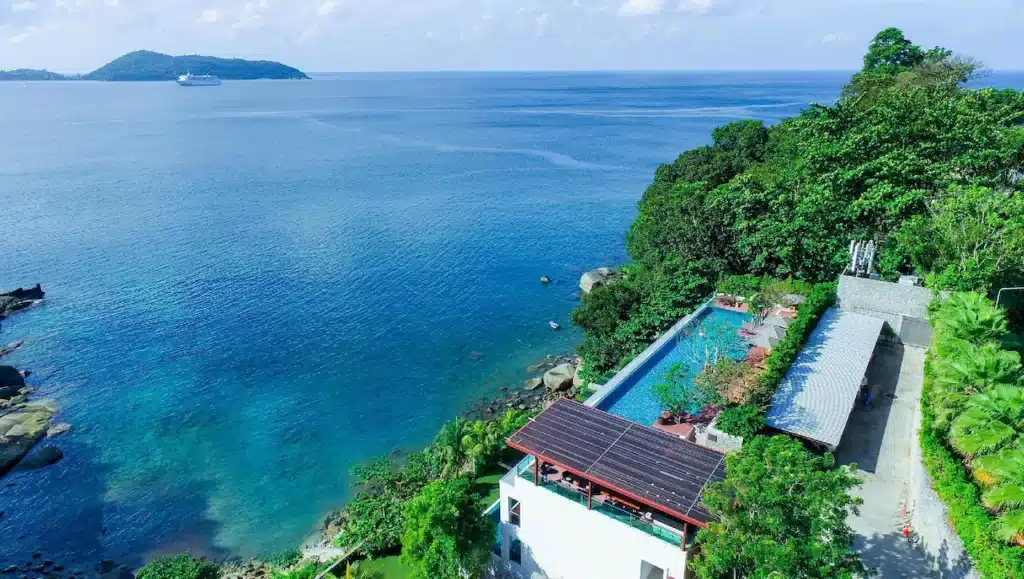 A stunning view of an infinity pool villa surrounded by lush greenery on a steep hillside overlooking a clear, blue ocean. A small building with a solar-paneled roof is adjacent to the Phuket pool villa. In the background, a cruise ship is sailing near a distant island under a partly cloudy sky.