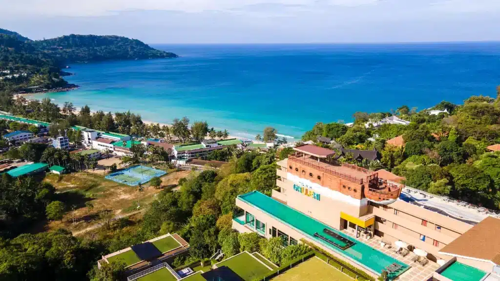 Aerial view of a coastal area with a mix of buildings and lush greenery. The turquoise ocean borders a sandy beach, curving around a forested peninsula. Prominent in the foreground is a multi-storey tan building with a green roof, one of the many hotels in Phuket surrounded by smaller structures and trees.