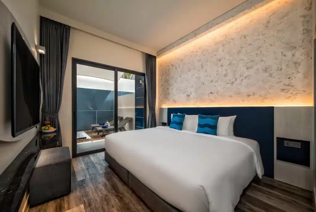 A modern hotel room at one of the premier hotels in Phuket, featuring a large bed adorned with white linens and blue accent pillows. A textured wall with soft backlighting sits behind the bed. To the left, a flat-screen TV is mounted on the wall. The room has a sliding glass door leading to a balcony with outdoor furniture.