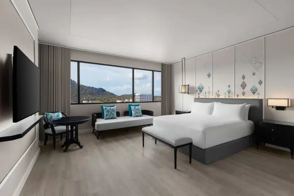 A modern, spacious hotel room in one of the finest hotels in Phuket, featuring a large bed with a white duvet and pillows. There is a white sofa with turquoise cushions, a small round table with chairs, a flat-screen TV mounted on the wall, and a window offering views of mountains and buildings. The decor is minimal and elegant.