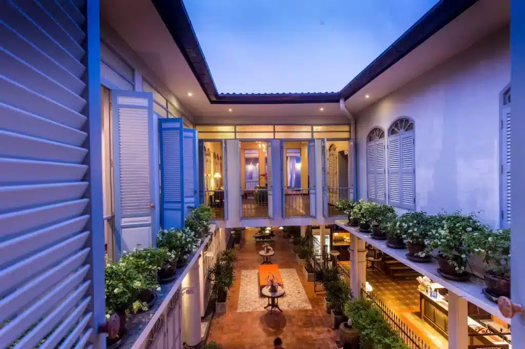 A serene courtyard of a two-story building with an open roof, featuring blue-shuttered windows and doors. The first floor has a pathway lined with potted plants, leading to a cozy outdoor seating area with a fire pit in the center. This hidden gem is perfect for those seeking off-the-beaten-path Phuket attractions.