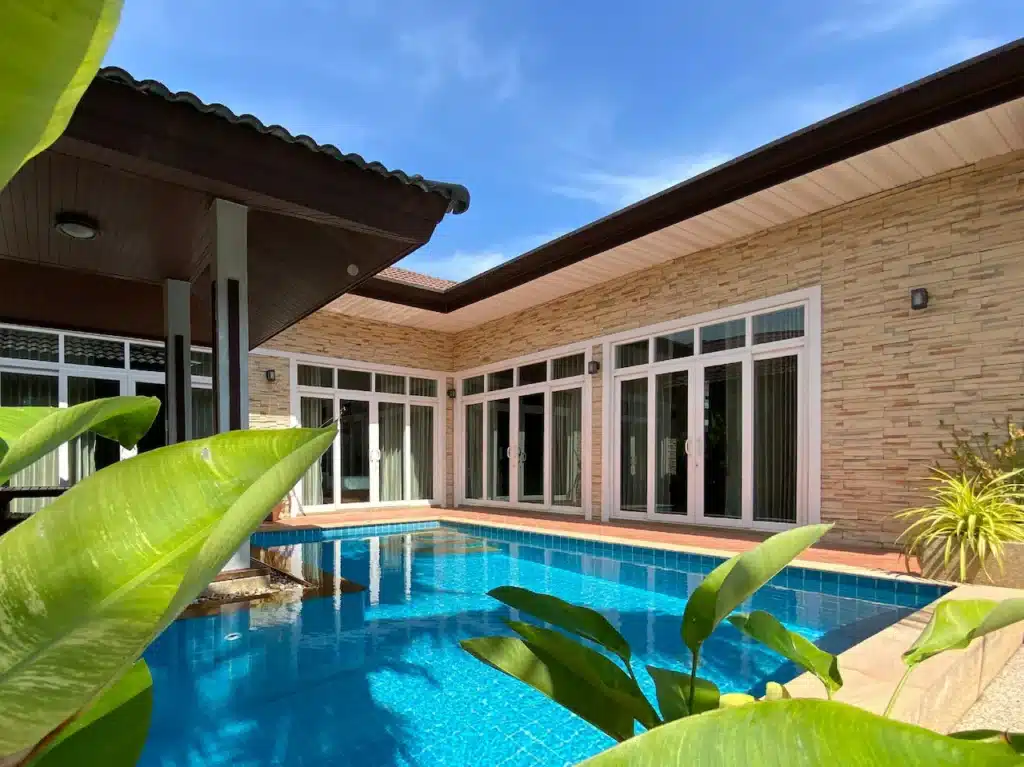 A modern Phuket pool villa with large sliding glass doors and a beige stone exterior surrounds a vibrant blue rectangular pool. Tropical green plants frame the foreground, while a clear blue sky stretches above, enhancing the serene atmosphere. The house features a covered patio section with a dark wood ceiling.