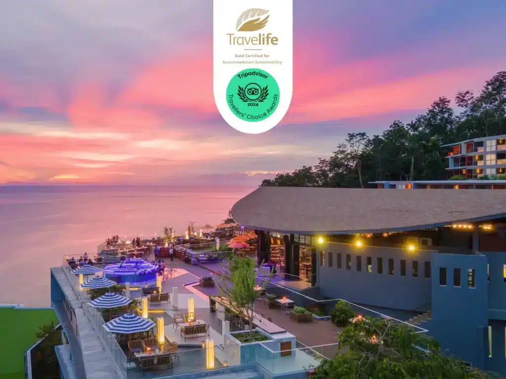 A luxurious oceanfront resort in Phuket is displayed at sunset, featuring infinity pools and outdoor seating areas with striped umbrellas. Above the resort is a circular green "Travelife" certification badge indicating environmental sustainability and a TripAdvisor "Travelers' Choice" award for 2024.