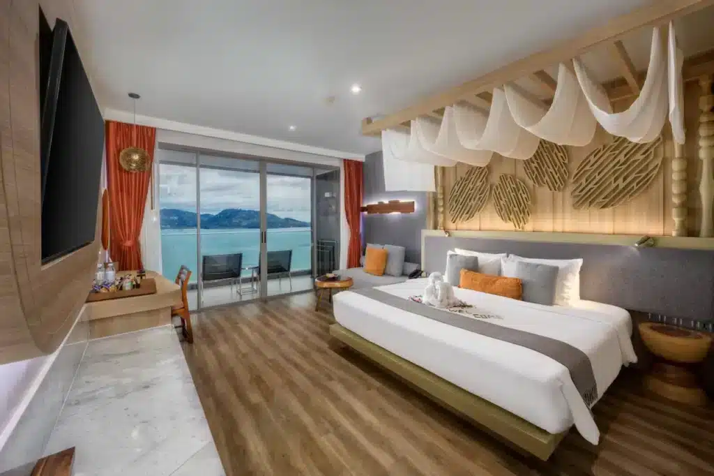 A modern hotel room with a large bed adorned with white linens and orange accent pillows. There is a TV on a wooden panel, a desk with a chair, and a balcony with glass doors offering an ocean view. Featuring wood flooring, a canopy above the bed, and unique wall art, this is one of the finest hotels in Phuke.