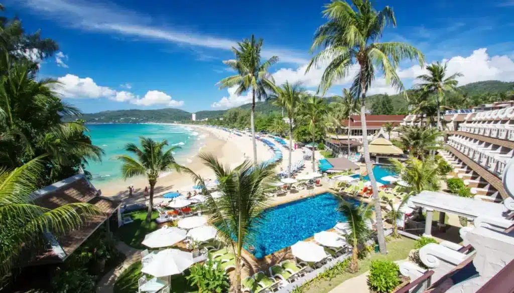 A tropical beach resort scene in Phuket features a large, vibrant blue pool surrounded by sun loungers and umbrellas. Palm trees are scattered throughout the resort area. The beach, lined with more umbrellas and visitors, stretches alongside the turquoise ocean. Lush green hills and a bright blue sky with scattered clouds complete the picturesque view.