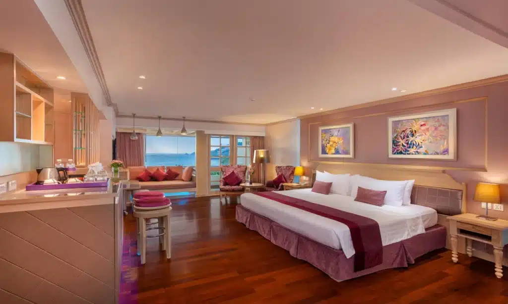 A spacious hotel room with a large bed adorned with white and maroon linens and matching pillows. The room features wooden floors, a seating area with an ocean view through large windows, warm lighting from table lamps on both sides of the bed, and artwork on pastel-colored walls—ideal for those seeking luxurious Private Pool Villas in Phuket.