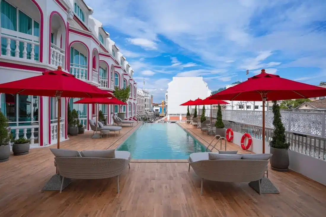 A serene outdoor pool area, featured in many Phuket travel guides, is flanked by a modern, pink-and-white building. Deck chairs and woven loungers are positioned on wooden decking, shaded by red umbrellas. The long, narrow pool is bordered by small trees and safety rings under a partly cloudy sky with blue patches.