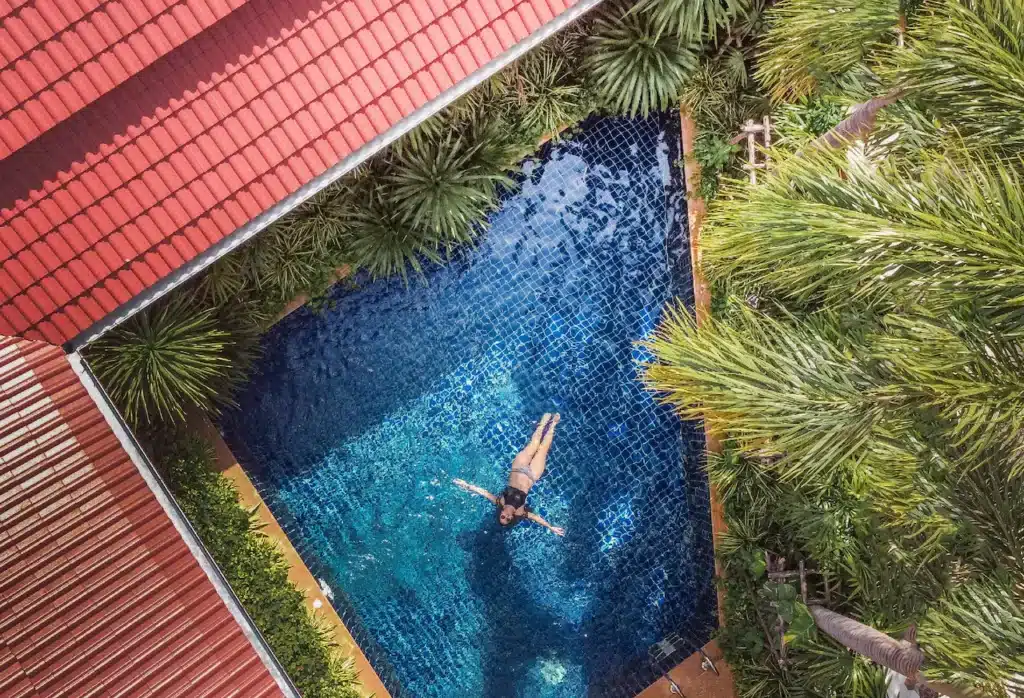 A person floats in a blue-tiled swimming pool surrounded by lush green foliage and red-roofed buildings. The aerial view of this Phuket pool villa showcases the contrast between the pool's vibrant blue and the greenery's rich tones. The person is centered and appears relaxed, enjoying the tranquil setting.