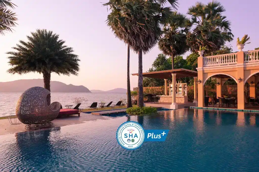 A luxurious seaside pool area at sunset, featuring an elegant, palm-tree-adorned landscape. The pool, part of one of the finest hotels in Phuket, is adjacent to a large, arched veranda. A shell-shaped lounge chair is placed by the water's edge. A certification badge with "SHA Plus+" is visible in the center of the image.