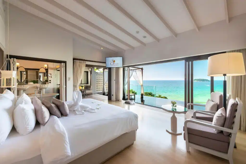 Luxurious beachside hotel room with a stunning ocean view. The room features a king-sized bed with white linens, plush pillows, modern furnishings, a TV, and a spacious open layout. Large sliding glass doors open to a balcony with lounge chairs, overlooking the Phuket pool villa’s turquoise sea and clear sky.