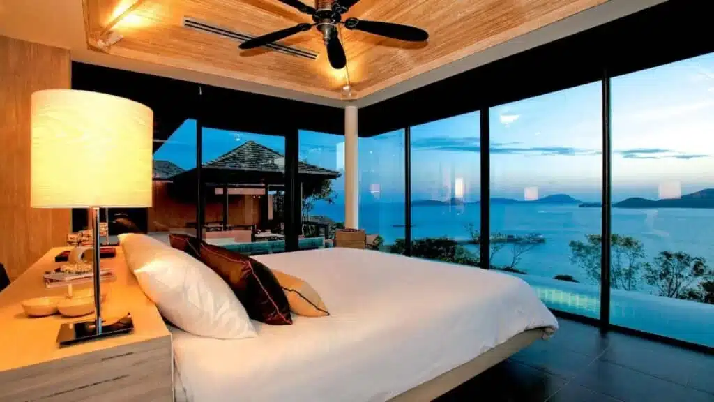 Luxurious bedroom with a large bed, bedside table, and lamp. The room features floor-to-ceiling windows offering a stunning view of an infinity pool, a patio, and a scenic ocean landscape with islands in the distance—typical of the best hotels in Phuket. A ceiling fan and modern decor complete the serene ambiance.