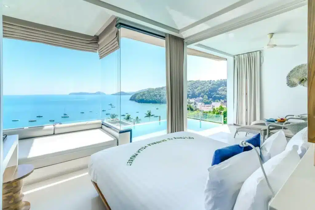 A bright and airy hotel room with large floor-to-ceiling windows offering a stunning view of a clear blue sea and green hillside. Located among the top hotels in Phuket, the room features a neatly made white bed, a cushioned window seat, modern furnishings, and a ceiling fan. Elegant, neutral curtains frame the windows.