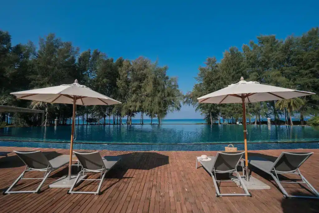 A serene scene featuring two lounge chairs under white umbrellas on a wooden deck by an infinity pool at one of the finest hotels in Phuket. Beyond the pool, trees with lush green foliage stand against a clear blue sky, with glimpses of the sea visible in the background, creating a tranquil and inviting atmosphere.