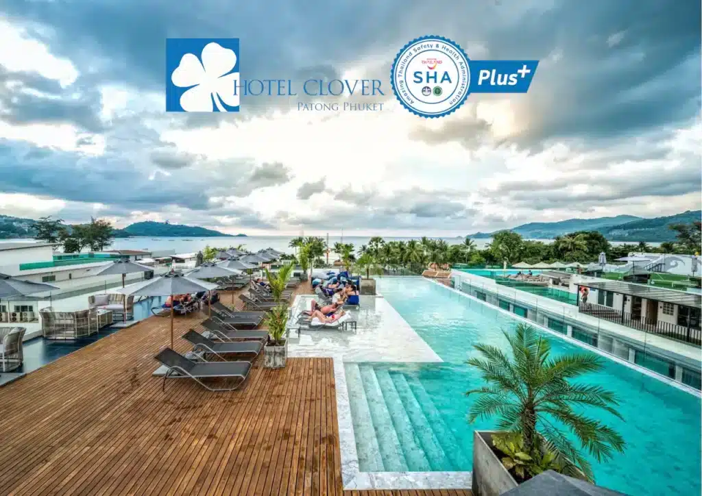 A rooftop infinity pool at Hotel Clover Patong Phuket, one of the top-rated hotels in Phuket, features wooden decking, sun loungers, and umbrellas. Guests are relaxing by the poolside with a view of lush greenery and distant mountains under a partly cloudy sky. The hotel's logo and SHA Plus+ certification badge are visible.