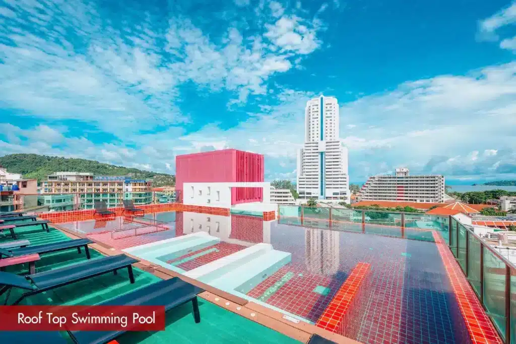 A rooftop swimming pool with clear blue water and red tiles offers a scenic view of Phuket's cityscape, with tall buildings and hills in the distance. Lounge chairs are arranged around the pool under a bright blue sky. A red sign in the bottom left corner reads "Rooftop Swimming Pool.