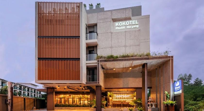 Modern hotel building with a sign reading "Kokotel Phuket Naiyang" on the facade, ideal for those seeking budget hotels near Phuket Airport. It features large wooden slats on the left, a glass entrance with warm lighting, and plants on the upper terrace. A lit outdoor seating area is visible through the glass. A blue sign is on the right side near the entrance.