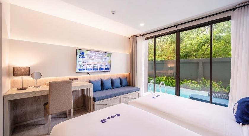 A modern hotel room featuring two neatly made beds with white linens and blue pillows. The room includes a desk, chair, and lamp on the left side. A flat-screen TV is mounted on the wall above a cushioned seating bench. Large sliding glass doors lead to a pool area surrounded by greenery, perfect for those seeking budget hotels near Phuket Airport.